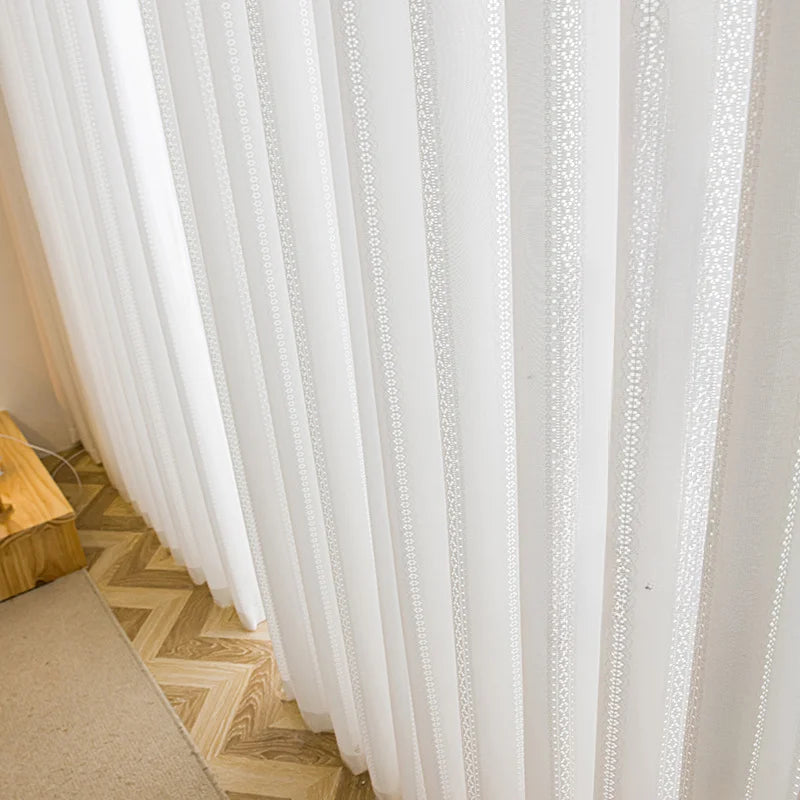 Rideaux en voile blanc brodé installés sur une grande baie vitrée, intérieur lumineux et naturel.