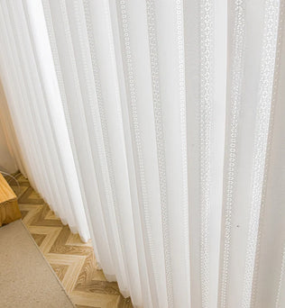 Rideaux en voile blanc brodé installés sur une grande baie vitrée, intérieur lumineux et naturel.