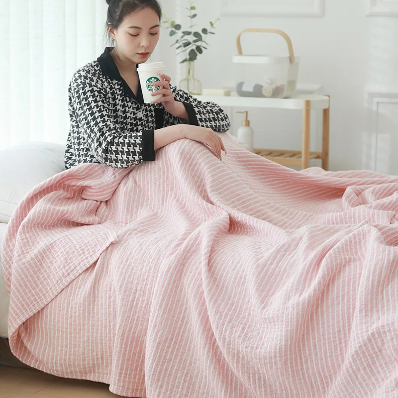 Une femme assise confortablement sous une couverture à rayures roses pendant qu'elle boit une boisson chaude dans une chambre lumineuse et accueillante.