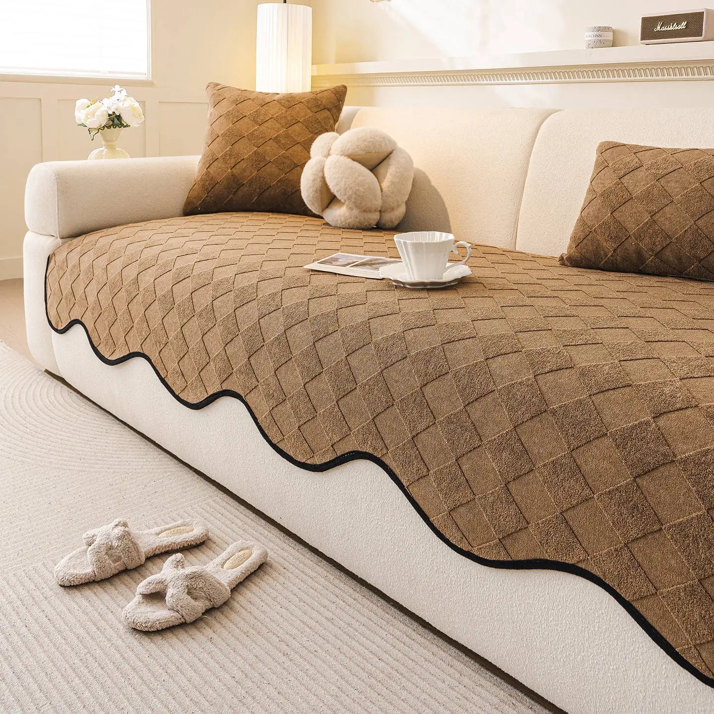 Couvrelit en peluche beige avec motif géométrique et ornements décoratifs, posé sur un canapé blanc dans une pièce confortable et chaleureuse.