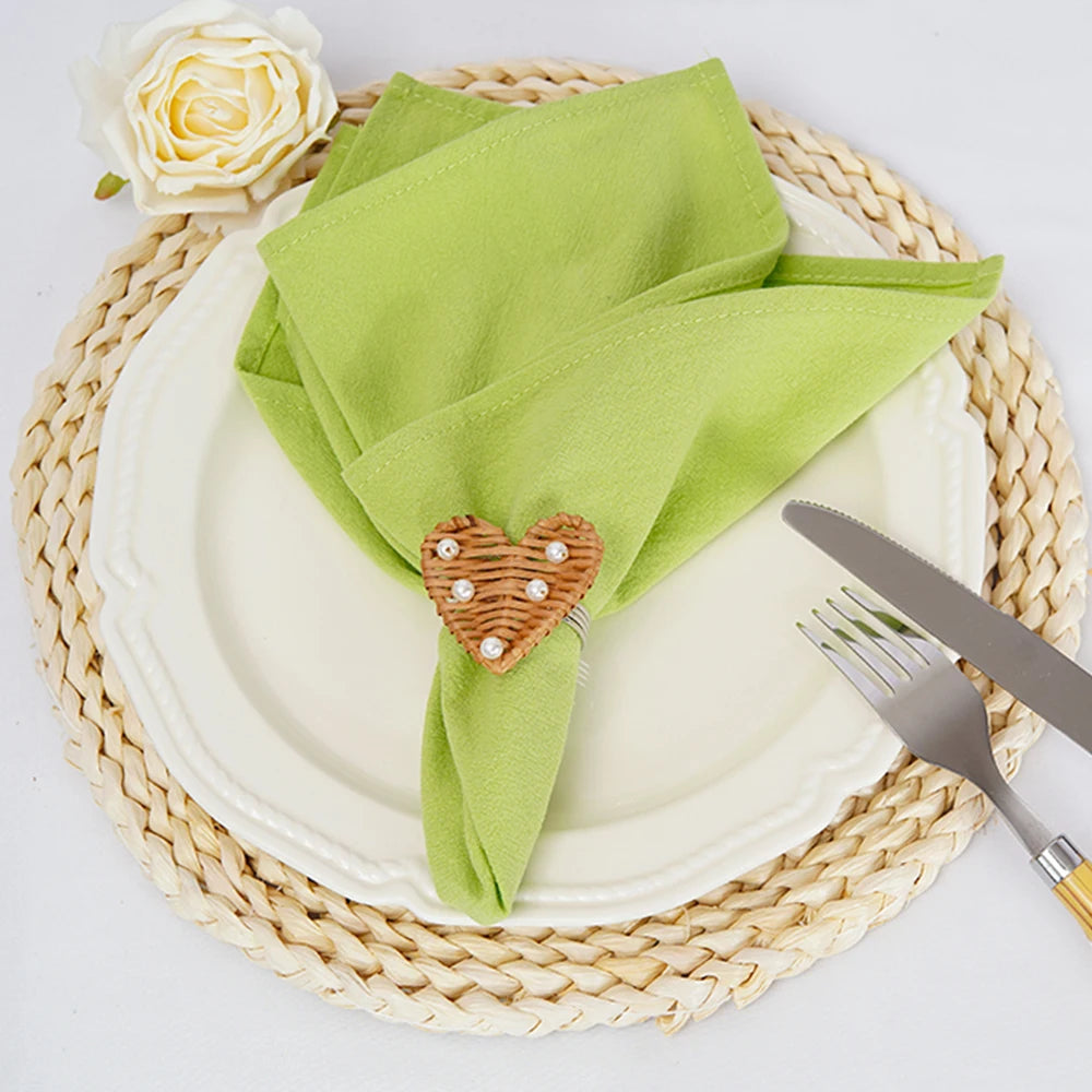 Une élégante serviette verte posée sur un tapis tressé, accompagnée d'une rose blanche et d'une décoration en forme de cœur en osier sur l'assiette, créant une ambiance harmonieuse et raffinée pour une table dressée.