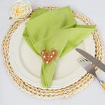 Une élégante serviette verte posée sur un tapis tressé, accompagnée d'une rose blanche et d'une décoration en forme de cœur en osier sur l'assiette, créant une ambiance harmonieuse et raffinée pour une table dressée.