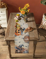 Table de cuisine en bois rustique avec un chemin de table orné de motifs d'Halloween et décoré de tournesols, de pommes et de pains, créant une atmosphère chaleureuse et automnale.