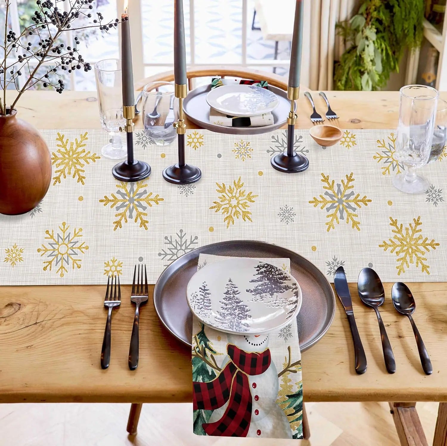 Une table festive prête pour un repas d'hiver, avec des assiettes décorées de motifs de flocons de neige dorés et gris, des chandeliers, des couverts et une nappe ornée de motifs similaires.