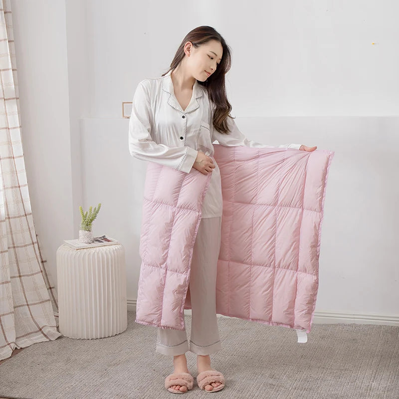 Une femme vêtue d'une chemise blanche et d'un pantalon rose pâle se tient debout dans une pièce avec un fauteuil et un rideau en arrière-plan, tenant une couverture rose dans ses mains.