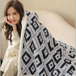 Une jeune femme souriante, habillée d'un pull blanc, enveloppée dans une couverture grise et blanche à motifs géométriques, profite d'un moment de détente et de confort.