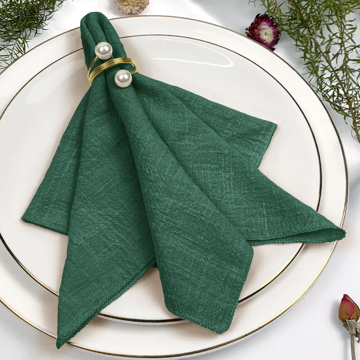 Un pli en tissu vert foncé avec des anneaux dorés et des perles sur un plat blanc disposé sur une table avec des fleurs décoratives.