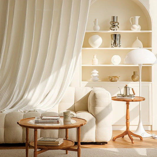 Voilage blanc effet froissé suspendu en arrière-plan d’un salon beige au mobilier organique et aux touches de bois naturel.