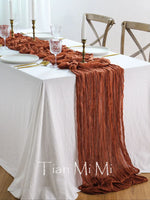 Une table élégante avec une nappe blanche, une chemin de table froissé de couleur rougeâtre, des bougies et des verres sur la table.