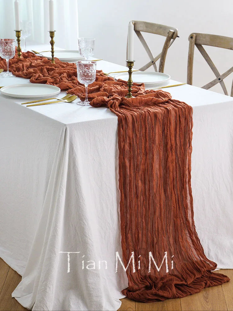 Une table élégante avec une nappe blanche, une chemin de table froissé de couleur rougeâtre, des bougies et des verres sur la table.