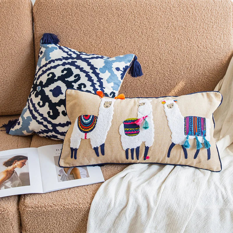 Un coussin rectangulaire avec des motifs de lamas décoratifs et des glands est posé sur un canapé beige, accompagné d'un magazine.