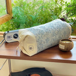 Un coussin de meditation cylindrique recouvert d'un motif paisible, posé sur une table en bois près d'une radio-réveil et d'une bougie dans un bol en bois, avec des plantes vertes en arrière-plan.