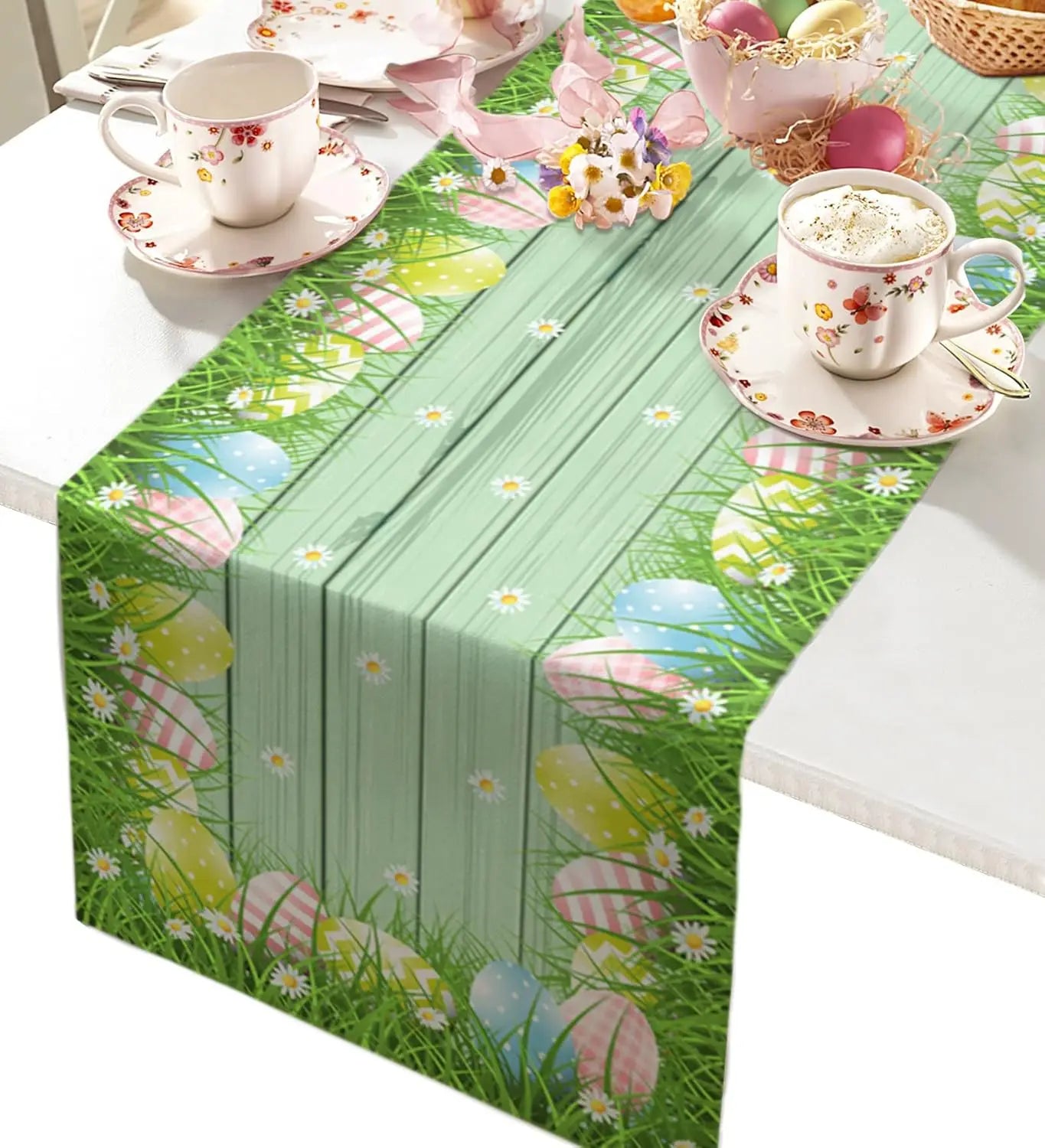 Une table de salle à manger printanière couverte d'un motif de fleurs, d'œufs de Pâques et d'herbe verte, accompagnée de tasses et de soucoupes ornées de motifs floraux.