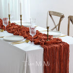 Une table élégante avec une nappe blanche, des bougies, des verres de cristal et une décoration de tissu froissé en couleur rouille, créant une atmosphère chaleureuse et raffinée.