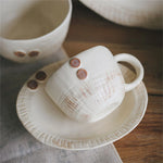 Deux tasses de thé en céramique blanche décorées de motifs géométriques et de points rouges, posées sur une table en bois.