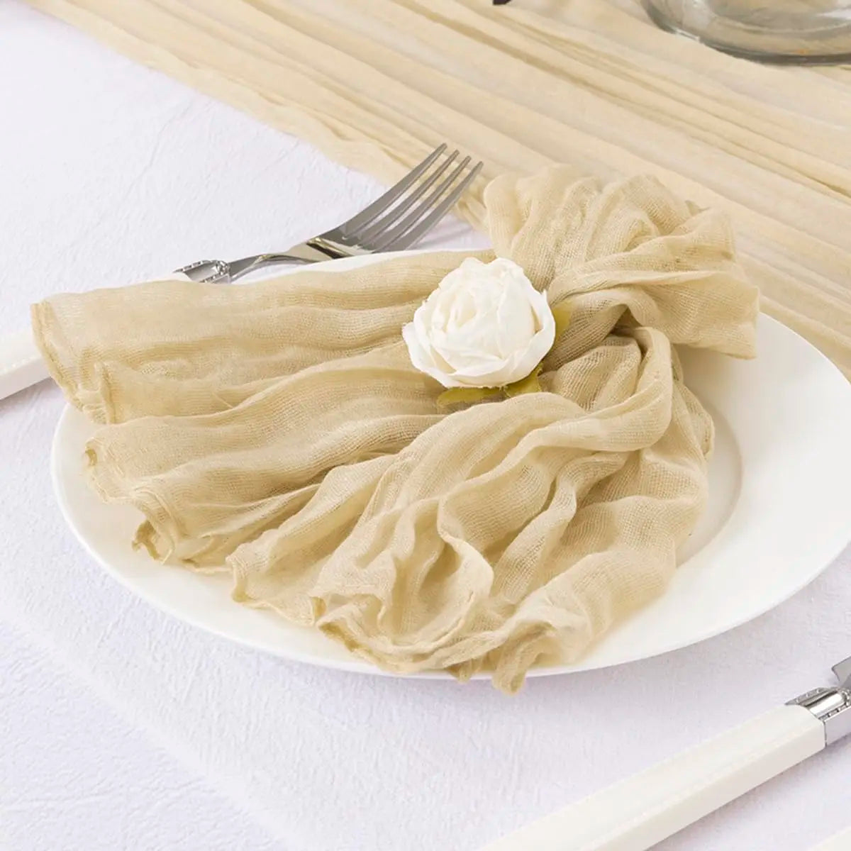 Assiette blanche avec une serviette en tissu beige drapée et décorée d'une rose blanche, accompagnée d'une fourchette et d'un couteau.