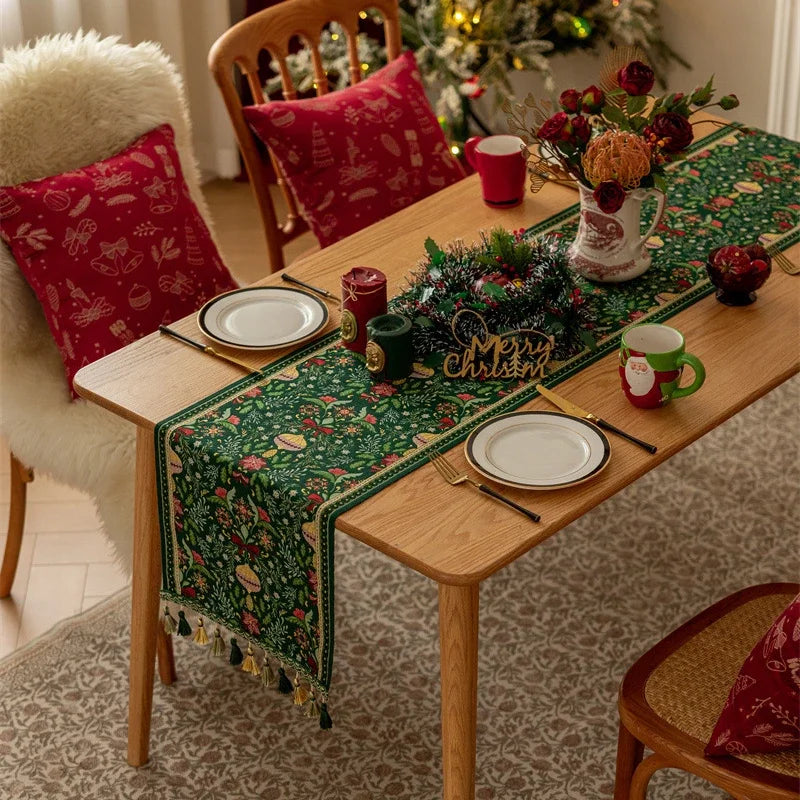 Une table élégamment décorée avec des décorations de Noël, comprenant une nappe verte ornée de motifs floraux, des coussins rouges décoratifs, des bougies et un vase avec des fleurs rouges, le tout encadré par un arbre de Noël illuminé.