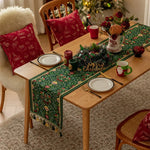 Une table élégamment décorée avec des décorations de Noël, comprenant une nappe verte ornée de motifs floraux, des coussins rouges décoratifs, des bougies et un vase avec des fleurs rouges, le tout encadré par un arbre de Noël illuminé.