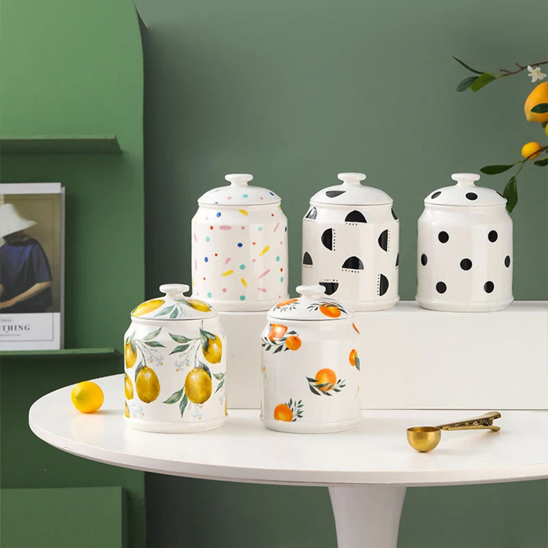 Plusieurs boîtes de rangement en céramique blanche décorées de motifs graphiques et de formes colorées posées sur une table ronde blanche, dans un décor vert foncé.