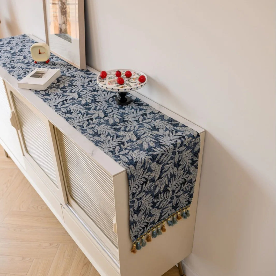 Chemin de table en jacquard bleu marine avec franges moutarde, sur un meuble blanc près d'un réveil rouge.