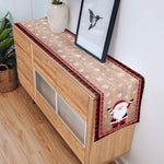 Meuble en bois clair avec un chemin de table motif flocons de neige et Père Noël, décoré pour Noël, avec un cadre mural et un vase décoratif.
