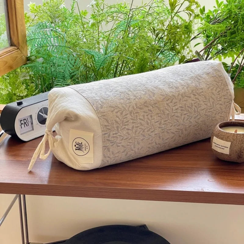 Un oreiller en forme de rouleau avec un motif discret posé sur une table en bois, accompagné d'un réveil et d'une bougie, devant une fenêtre donnant sur un paysage verdoyant.