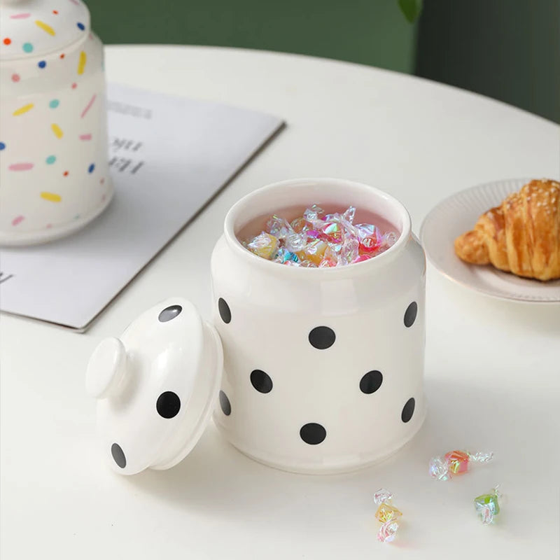 Une tasse en céramique blanche aux motifs de points noirs, remplie de bonbons colorés, accompagnée d'un croissant et d'un livre ouvert sur une table blanche.