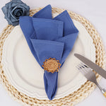 Sur une assiette blanche repose un arrangement de serviettes de table bleues pliées, ornées d'un motif floral en bois. Une fourchette et un couteau sont disposés à côté.