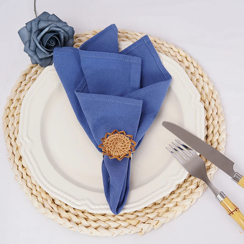 Sur une assiette blanche, des serviettes de table bleu marine sont pliées de manière décorative avec une rose grise posée dessus, entourées de couverts métalliques sur un dessous de plat tressé.
