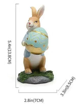 Figurine en porcelaine d'un lapin de Pâques tenant un œuf de Pâques décoré assis sur l'herbe.