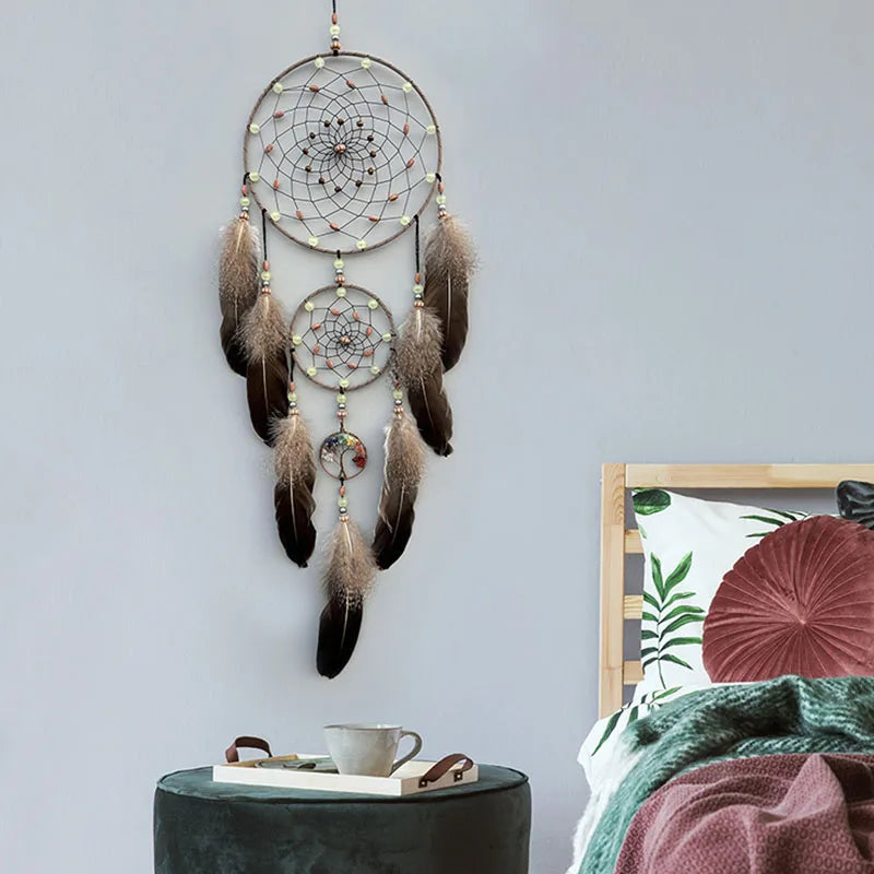 Un attrape-rêves orné de plumes brunes et vertes, avec des motifs circulaires décoratifs, suspendu sur un mur avec des accessoires de chambre à l'arrière-plan.