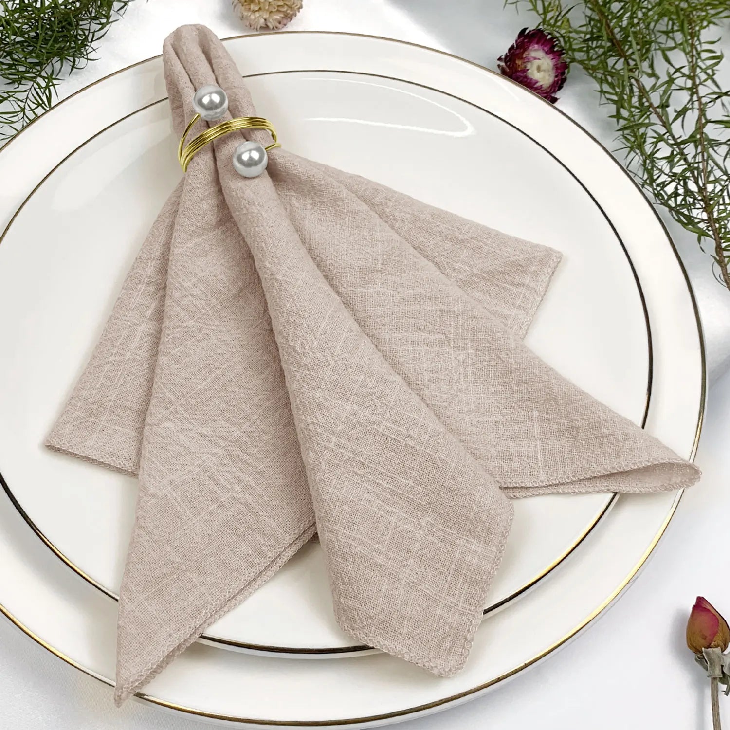 L'image montre une assiette blanche avec des serviettes de table beiges pliées de manière artistique, ornées de perles décoratives. Un beau bouquet de fleurs roses accompagne cette mise en scène élégante.