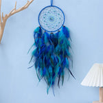 Un attrape-rêves bleu et vert avec un motif spiralé, orné de plumes bleues et vertes, suspendu à un bâton en bois sur un mur.