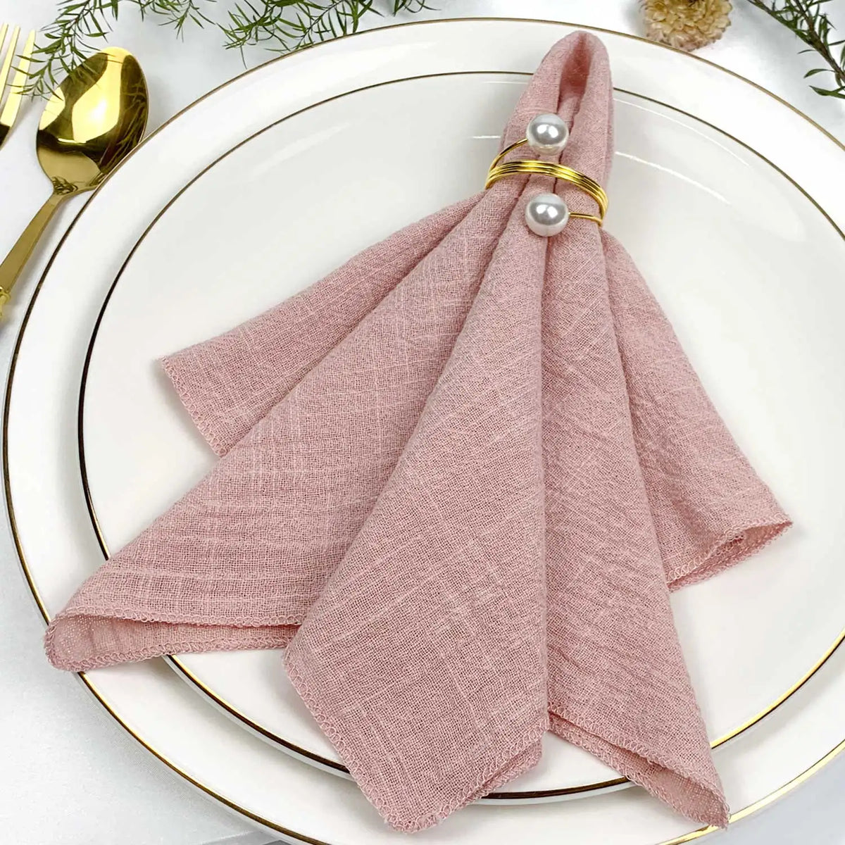 Une nappe de table blanche avec des décorations végétales et des couverts dorés, sur laquelle repose une serviette de table rose poudré ornée de perles.