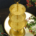Cet image montre un élégant plat à gâteaux doré à trois niveaux, orné de motifs floraux finement ciselés, posé sur une surface blanche et accompagné de décorations végétales, créant une atmosphère festive et raffinée.