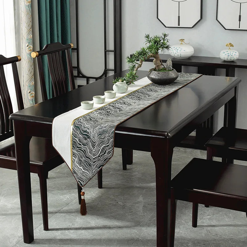 Cette image présente une table à manger élégante avec un chemin de table en motif abstrait noir et blanc, ornée de plantes en pot et de vases en céramique, dans un intérieur moderne avec des rideaux en velours vert.
