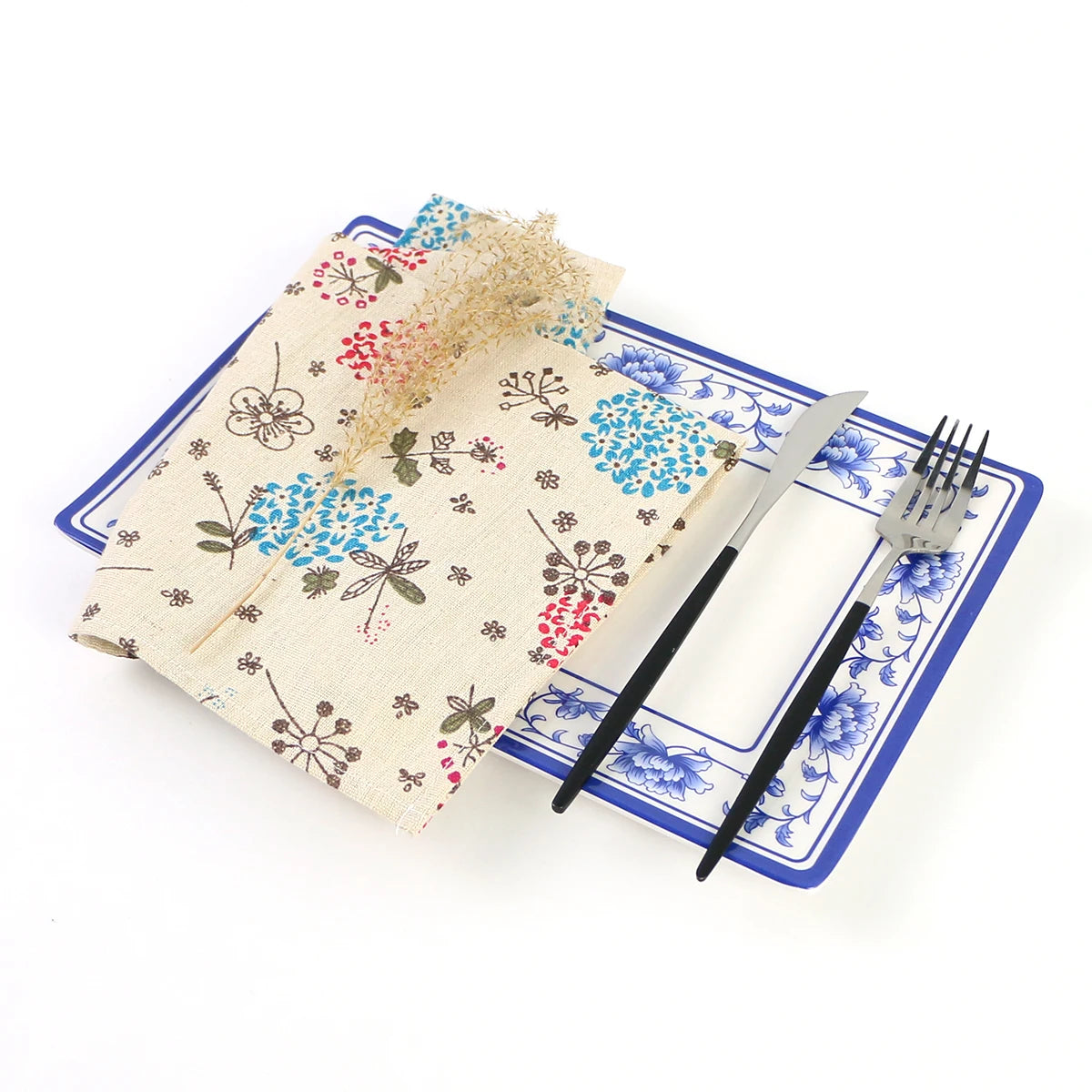 Cette image montre un set de table en tissu beige orné de motifs floraux colorés, accompagné de couverts sur un fond bleu clair.