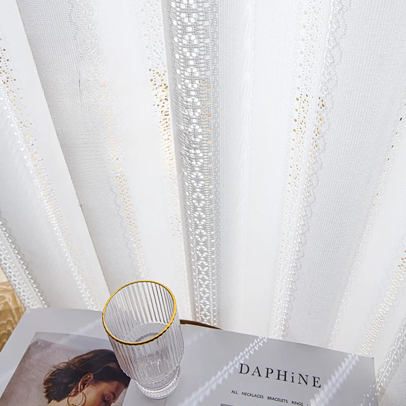 Détail du voilage blanc à ruflette avec motifs brodés et un verre posé sur une table à côté.