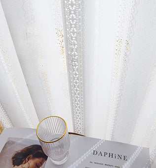 Détail du voilage blanc à ruflette avec motifs brodés et un verre posé sur une table à côté.