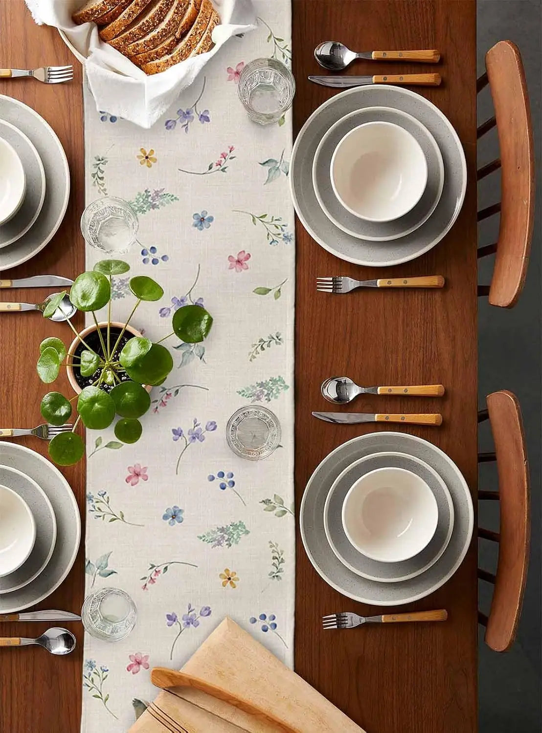 L'image montre une table de salle à manger avec une nappe fleurie, des assiettes blanches, des couverts en bois et un petit arrangement floral en pot. L'ensemble crée une atmosphère chaleureuse et conviviale.