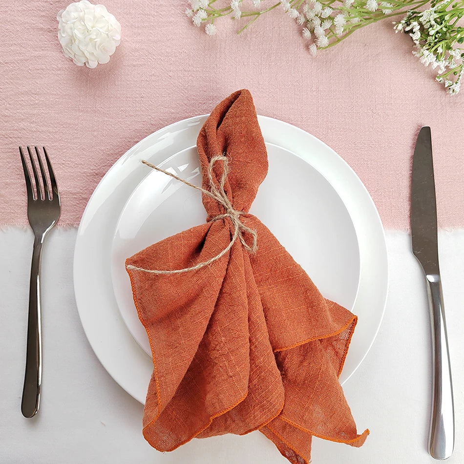 L'image montre une assiette blanche avec une serviette de table en lin orange, pliée de manière décorative et attachée avec une corde. L'assiette est entourée de fleurs blanches et de fourchettes et couteaux en métal sombre.