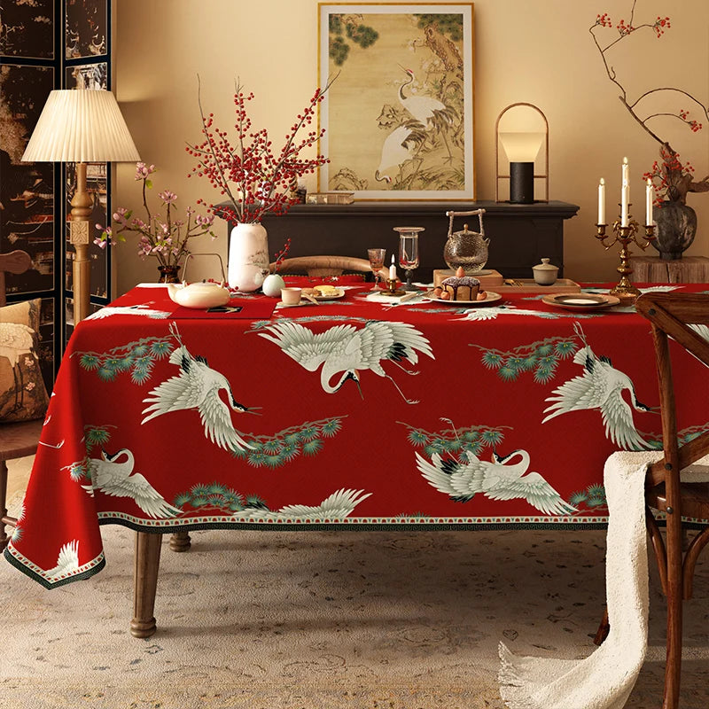 Cette image présente une table de salle à manger élégamment dressée, avec un magnifique motif de grues et de branches de pin sur une nappe rouge, entourée d'accessoires décoratifs dans un intérieur chaleureux et harmonieux.