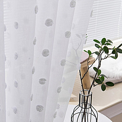 Voilage blanc à pois en relief, illuminé par la lumière naturelle d’une pièce cosy, décorée d’un vase et d’une plante verte.