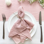 Une élégante mise en place avec une nappe rose poudrée, une jolie serviette nouée et des couverts en métal sur une assiette blanche.