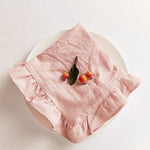 Cette image montre un gros plan d'un plateau blanc avec un linge de table rose froissé, sur lequel se trouvent quelques petites pommes rouges attachées par une branche.