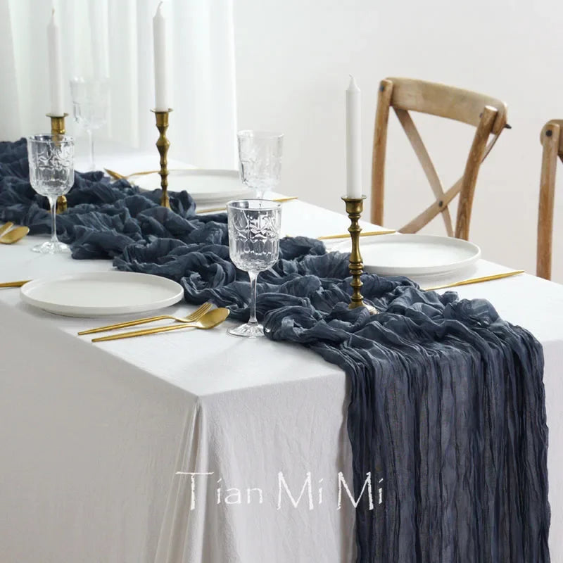 La table est soigneusement mise avec des assiettes blanches, des verres de cristal, des couverts dorés et une nappe blanche ornée d'un chemin de table en satin gris foncé, créant une ambiance élégante et raffinée.
