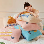 Une femme assise sur un lit, entourée de plusieurs coussins aux motifs et couleurs pastels, semble se reposer confortablement.