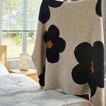 Couverture douce avec imprimé fleuri noir et beige sur fond clair, posée sur un lit près d'une fenêtre.