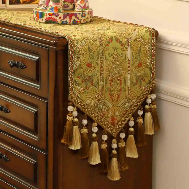 Chemin de table décoratif avec des motifs floraux et ornementaux dorés, des pompons et des glands formant une bordure élégante au-dessus d'un meuble en bois foncé.