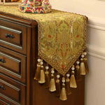 Chemin de table décoratif avec des motifs floraux et ornementaux dorés, des pompons et des glands formant une bordure élégante au-dessus d'un meuble en bois foncé.